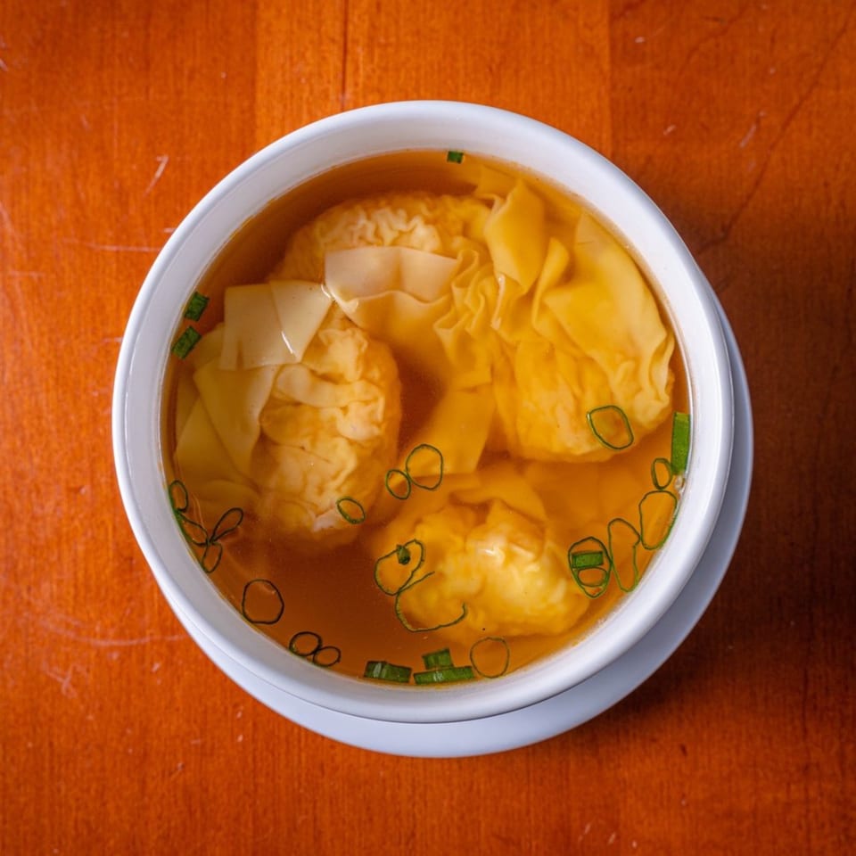 A white bowl of Modern Fusion wonton soup with clear broth and green onions on a wooden table.
